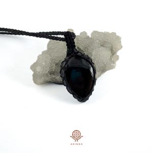 May include: A black gemstone pendant, teardrop-shaped, set in a woven black cord. The pendant is suspended from a matching black cord necklace. The background is a light grey, textured surface. The brand name "ARINNA" is at the bottom.