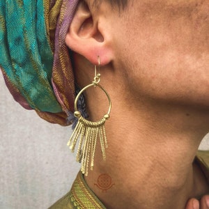 Peut inclure: Boucles d'oreilles créoles dorées avec une frange de brins pendants. Les boucles d'oreilles sont portées par une personne avec un foulard coloré.