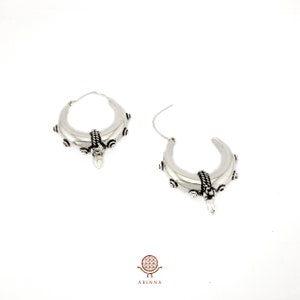 May include: A pair of silver hoop earrings with a crescent moon shape and intricate detailing. The earrings have a textured surface and a small, pointed pendant hanging from the bottom.
