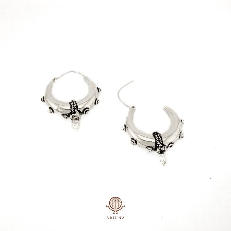 May include: A pair of silver hoop earrings with a crescent moon shape and intricate detailing. The earrings have a textured surface and a small, pointed pendant hanging from the bottom.