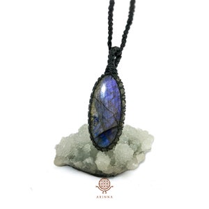 May include: A black cord necklace with a blue and grey oval labradorite gemstone pendant. The pendant is wrapped in black cord and is sitting on a white rock.