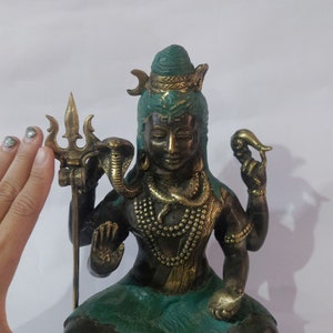 May include: Bronze statue of a seated deity with a trident, snake, and other symbolic items. The statue has a green and gold color scheme, with intricate details and a serene expression. The statue is approximately 8 inches tall.