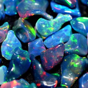 Könnte beinhalten: Eine Sammlung polierter, unregelmäßiger Opal-Edelsteine mit einer tiefblauen Grundfarbe und lebhaften Blitzen in Neongrün, leuchtendem Rot und elektrischem Blau, die über die glatten, durchscheinenden Oberflächen jedes Steins spielen.