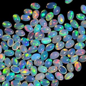 Könnte beinhalten: Eine Sammlung zahlreicher kleiner, oval geformter Cabochon-Opale. Jeder Edelstein weist eine durchscheinende, milchig-weiße Basis mit lebhaften, irisierenden Farbblitzen in Grün, Blau, Orange und Pink auf der Oberfläche auf.