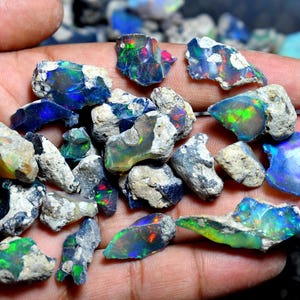 Könnte beinhalten: Eine Sammlung roher, unpolierter Opalsteine mit irisierenden Blitzen in Blau, Grün und Rot auf einer dunklen, felsigen Matrix. Diese natürlichen Edelsteine weisen unterschiedliche Formen und Texturen auf und zeigen ein lebhaftes Farbspiel.
