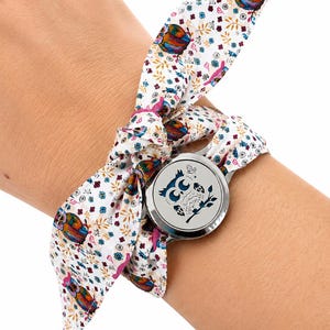 Peut inclure: Une montre-bracelet dotée d'un boîtier rond argenté avec un motif de hibou bleu foncé sur le cadran. Le bracelet est constitué d'un foulard en tissu blanc à motifs de hiboux colorés, de petites fleurs et de feuillages, noué au poignet.