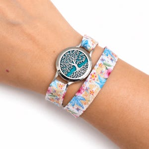 Puede incluir: Una pulsera envolvente con un medallón circular plateado con un diseño de árbol de la vida recortado sobre un fondo verde azulado. La banda es de tela con un estampado floral multicolor en tonos rosa, amarillo, azul y verde.