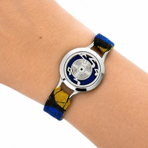 Peut inclure: Un bracelet composé d'une breloque circulaire en métal argenté avec un motif découpé représentant un disque vinyle et des notes de musique. La breloque est sur fond bleu foncé et fixée à un bracelet en tissu à motif bleu, jaune et noir.