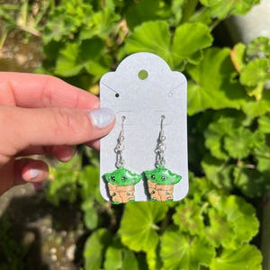 May include: A pair of silver dangle earrings with green and brown cartoon character charms. The charms are of a small green alien-like creature with large eyes.