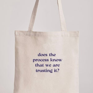Op de afbeelding: Een crèmekleurige draagtas met de tekst "does the process know that we are trusting it?" in donkerblauw. De tas heeft twee lange handvatten en is gemaakt van een stevige stof, geschikt voor het dragen van diverse items.
