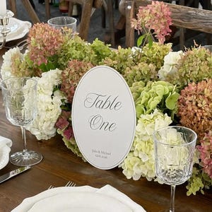 Peut inclure: Un panneau de table ovale avec le texte Table One, Julia & Michael, et 12.06.2026, placé parmi des hortensias blancs et roses sur une table en bois avec des assiettes blanches, des verres transparents, des couverts en argent et des serviettes blanches.