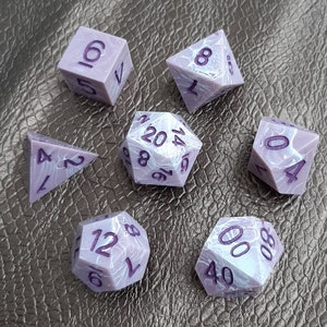 May include: Set of seven purple and white dice with black numbers. The dice are in various shapes, including a d4, d6, d8, d10, d12, and d20.
