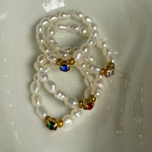 Op de afbeelding: Vier parelringen met gouden accenten en gekleurde edelstenen. De ringen zijn op elkaar gestapeld en liggen op een wit oppervlak.