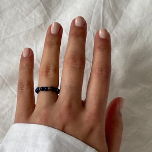 May include: A beaded ring featuring dark blue beads and a small gold accent bead. The ring is worn on a finger, with a white shirt sleeve in the background. The ring is a simple, elegant piece of jewelry.