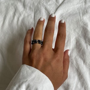 Peut inclure: Une bague en perles noires avec des accents dorés. La bague est portée sur une main avec du vernis à ongles blanc.