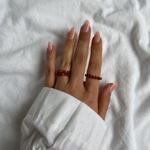 May include: Two red beaded rings on a hand with nude nail polish.