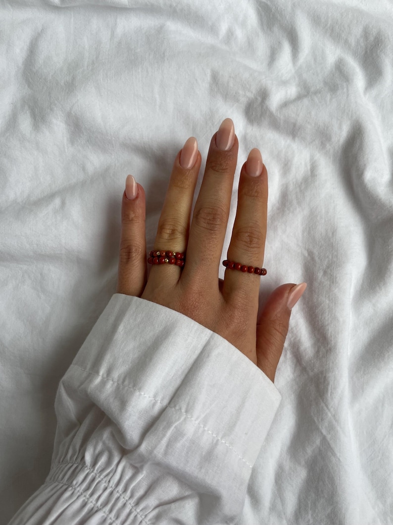 May include: Two red beaded rings on a hand with nude nail polish.