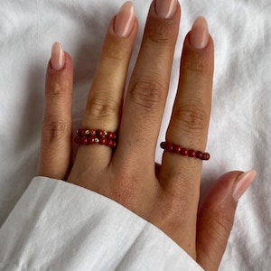 Op de afbeelding: Twee rode kralenringen op een hand met nude nagellak. De ringen zijn gemaakt van kleine, ronde kralen en zijn op elkaar gestapeld.