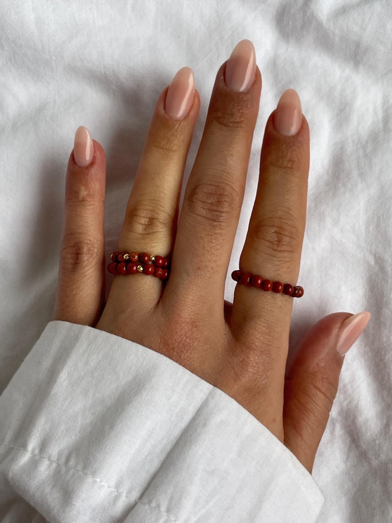 May include: Two red beaded rings on a hand with nude nail polish. The rings are made of small, round beads and are stacked on top of each other.