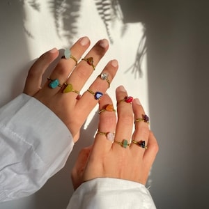 May include: A set of gold rings with different colored stones. The stones are in various shapes and sizes, including round, oval, and triangular. The rings are stacked on a person's fingers.