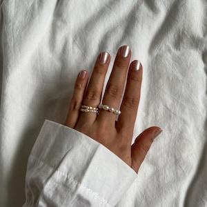 Könnte beinhalten: Zwei zarte Perlenringe an einer Hand mit hellrosa Nagellack. Die Ringe bestehen aus kleinen, weißen Perlen und sind übereinander gelegt.