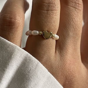 Puede incluir: Un anillo de oro con un dije de corazón y cuentas de perlas blancas.