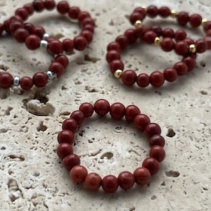 May include: Four red gemstone beaded rings on a light beige textured surface. The rings are made of small, round beads.