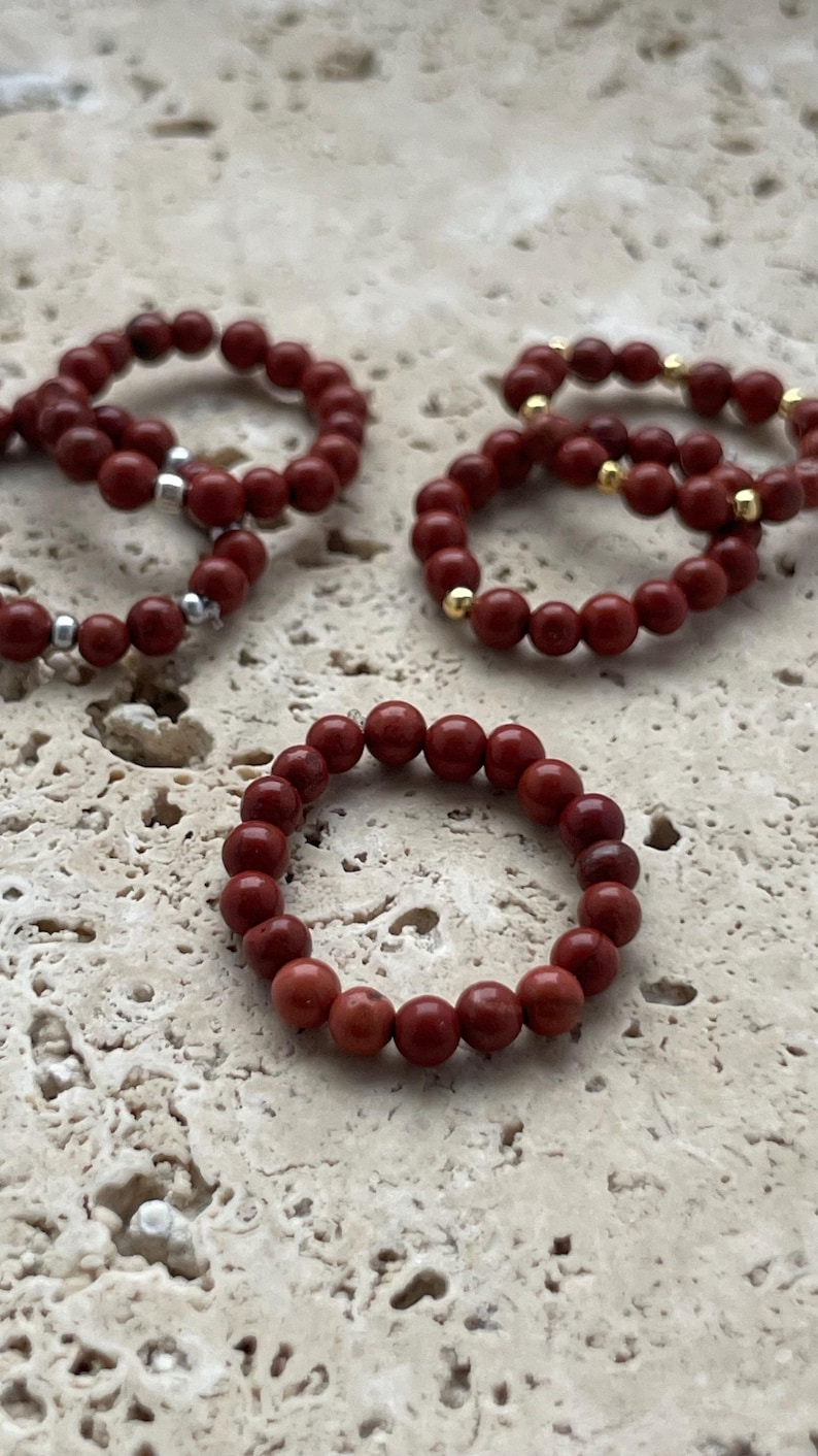 May include: Four red gemstone beaded rings on a light beige textured surface. The rings are made of small, round beads.