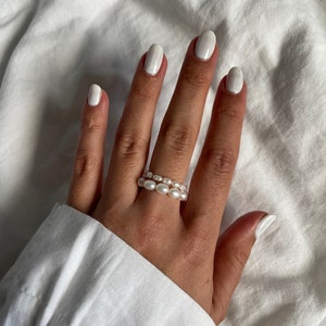 Könnte beinhalten: Nahaufnahme einer Hand, die zwei Perlenringe trägt. Die Ringe bestehen aus kleinen, weißen Perlen und sind übereinander gestapelt. Die Hand trägt weißen Nagellack.