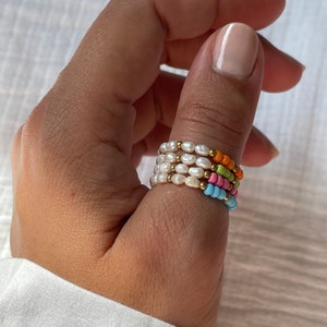 Op de afbeelding: Een set van vier kralenringen gestapeld op een vinger. De ringen zijn gemaakt van witte parels en gekleurde kralen in tinten oranje, roze, groen en blauw.