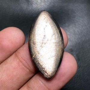 以下が含まれることがあります： メタリックな金茶色の光沢を持つ、研磨された細長い宝石。石は滑らかな表面を持ち、色と質感に微妙な変化があります。形状は対称的で、両端が尖っています。宝石は手で持たれています。