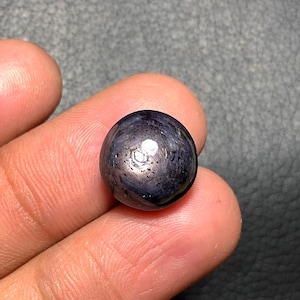 May include: A dark blue, spherical gemstone with a polished surface. The gem has a reflective, almost metallic sheen, with lighter areas highlighting its round shape. The stone is held in a person's hand, showcasing its size and detail.
