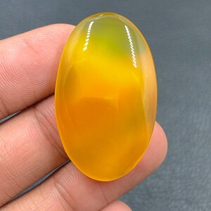Puede incluir: Un cabujón de ágata ovalado liso, amarillo y verde. La piedra tiene una apariencia translúcida y vidriosa.
