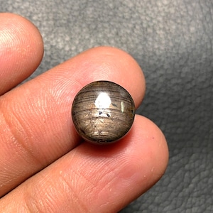 May include: A round, polished gemstone with a dark brown and gold striped pattern. The gemstone is held between fingers, showcasing its spherical shape and reflective surface. The stone appears to be a cabochon, suitable for jewelry.