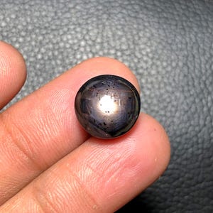 May include: A dark, round gemstone with a polished surface. The gem has a deep, purplish-black color with a bright, reflective spot in the center. The stone is held between two fingers, showcasing its size and shape.