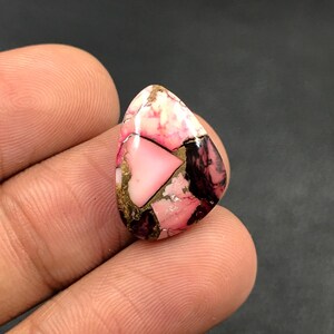 Peut inclure: Une pierre précieuse en forme de goutte d'eau avec une mosaïque de couleurs rose, noire et dorée. La pierre présente un triangle rose central et un mélange de textures, créant un aspect unique. La pierre est tenue entre les doigts.