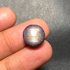 May include: A round, polished gemstone with a dark purple hue and a subtle, golden sheen. The surface displays fine, linear striations, creating a unique visual texture. The gem is held between fingers, showcasing its size and shape.