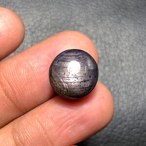 May include: A polished, spherical gemstone with a dark blue and gray color scheme. The gem has a reflective, white spot and visible striations across its surface. The stone is held between two fingers.