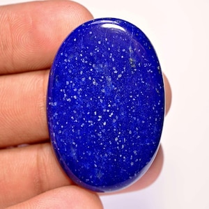 Puede incluir: Gema ovalada de lapislázuli pulido. La piedra azul oscuro está salpicada de pequeñas motas blancas, creando una apariencia estrellada. La gema se sostiene en una mano, mostrando su superficie lisa y brillante.