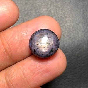 May include: A round, polished gemstone with a star effect. The gem is primarily a deep purple color with a shimmering, iridescent quality. The surface has a pattern of intersecting lines, creating a starburst effect. Small dark inclusions are visible.