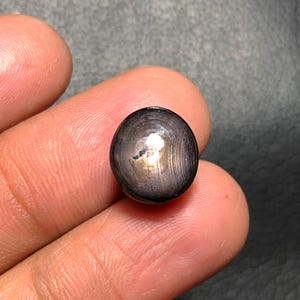 May include: A dark, spherical gemstone with a polished surface. The gem has a black base color with a star-like effect and a reflective sheen. The stone is held between fingers, showcasing its size and shape.