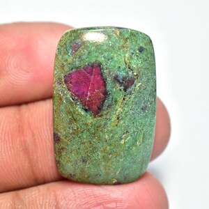 Peut inclure: Une pierre précieuse rectangulaire avec une couleur de base verte vibrante, parsemée de teintes vertes et brunes plus foncées. Une structure cristalline rouge foncé proéminente est centrée dans la pierre. La pierre est tenue par une main.