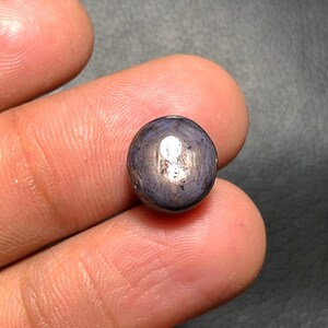 May include: A round, polished gemstone with a dark blue hue and a star-like pattern on its surface. The gem has a reflective, metallic sheen and appears to be held between fingers. The stone's surface has subtle variations in color and texture.