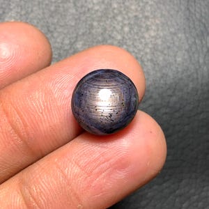 May include: A round, polished gemstone with a dark blue and silver color scheme. The stone has a reflective surface with visible striations and small dark inclusions. The gem is held between fingers, showcasing its size and shape.