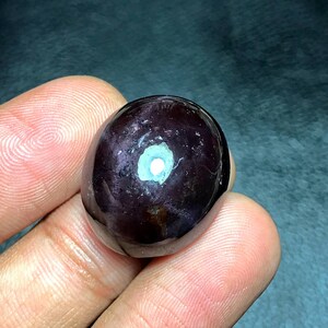 以下が含まれることがあります： 滑らかで丸みを帯びた形の、研磨された濃い紫色の宝石。石は光沢のある表面を持ち、明るい白と水色のインクルージョンがあります。宝石は手で持たれています。