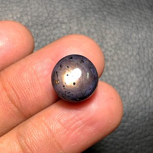 May include: A round, polished gemstone with a dark purple hue and a prominent star-like effect. The stone has a reflective, lustrous surface with a central, bright spot. The gem is held in a hand, showcasing its size and shape.