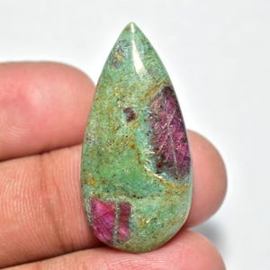 Peut inclure: Une pierre précieuse en forme de goutte d'eau, avec une base verte et des inclusions rouges vives. La pierre polie a une surface lisse et un aspect naturel. La pierre est tenue dans une main, mettant en valeur sa taille et sa forme.