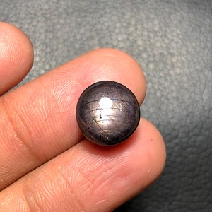 May include: A dark, spherical gemstone with a polished surface. The gem has a deep, almost black color with subtle streaks and a bright, reflective spot. The stone is held in a person's hand, showcasing its size and detail.