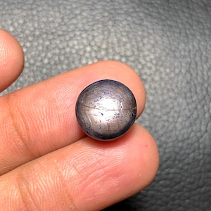 May include: A small, spherical gemstone with a dark, metallic appearance. The surface features a pattern of lines, resembling a miniature globe. The gem is held between two fingers, showcasing its size and detail.