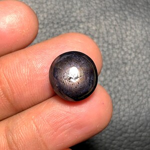 May include: A dark, spherical gemstone with a polished surface. The gem has a deep, iridescent color, with hints of purple and gold. The stone is smooth and reflective, catching the light. The overall appearance is of a high-quality, natural stone.